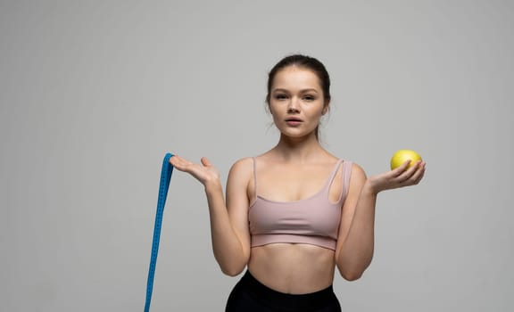 Confused attractive sexy brunette women in sport outfit standing with a measuring tape and holding a apple on a white background. Beauty and health concept. Healthy eating and diet