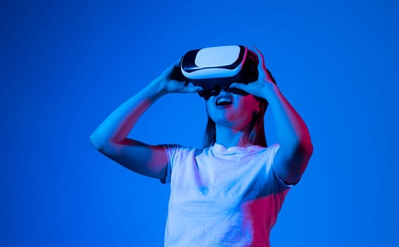 Metaverse concept of young brunette woman in white t-shirt wearing VR headset and playing video games and interacting with virtual reality in neon light