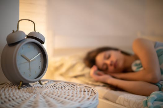 The girl in bed sleeps in the bedroom with an alarm clock on the cabinet next to her. The brunette has her eyes closed and smiles softly in her sleep.