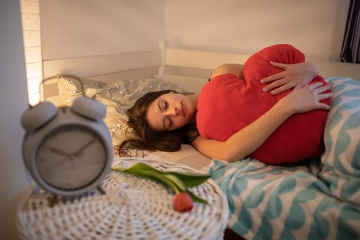 A girl sleeps with a red heart-shaped pillow. Bright bedroom with decorations. A large alarm clock stands on the bedside table.