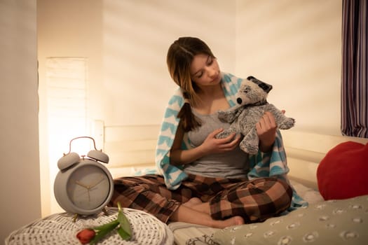 The girl holds the cuddly teddy bear in her hands. Bright bedroom with decorations. The light of a lamp on the wall.