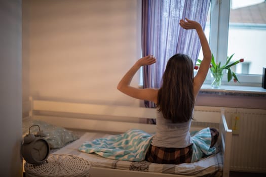 A rested woman stretches and looks out the window. Bright bedroom with decorations.
