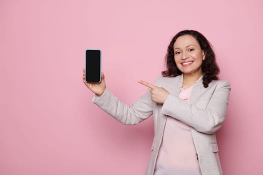 Middle-aged pretty woman in casual gray jacket, smiling looking at camera, showing smartphone with blank screen with free space for your mobile application or advertising, isolated on pink background