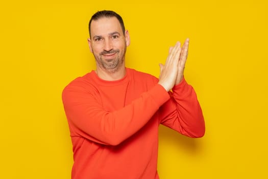 Bearded hispanic man wearing a red hoodie clapping excited for the great performance he just witnessed, isolated on yellow studio background