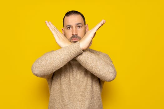 No way, absolutely not. Portrait of bearded hispanic man in turtleneck showing x sign with crossed hands meaning stop this is the end. Isolated on yellow studio background.