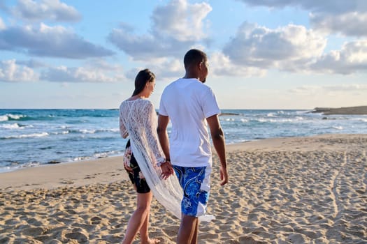 Young happy couple walking on the beach holding hands, back view. Honeymoon, dating, happiness, relationship, vacation, love, romance concept