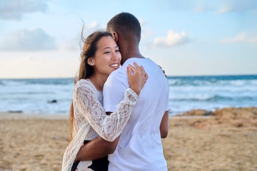 Young loving couple hugging on the beach. Face of beautiful Asian woman, man back. Love, relationship, honeymoon, vacation together, happiness, romantic, travel, family concept