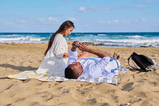 Young couple relaxing on the beach, lying sitting on the sand with smartphones. Summer autumn vacation together, lifestyle. technology, family concept