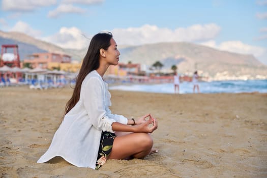 Young woman practicing yoga on beach. Beautiful Asian female meditating, sitting in lotus position on sand, on seashore. Beauty, lifestyle, meditation, relaxation, lifestyle, nature, people concept