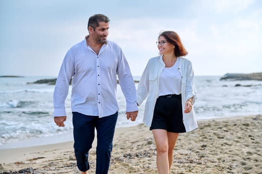 Happy middle-aged couple walking together on the beach. Mature man and woman holding hands walking along seashore. Relationship, lifestyle, vacation, tourism, sea nature, people concept