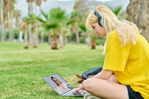 Remote online lecture on laptop screen, web chat with teacher individual consultation of teenage. College high school student girl sitting on grass in park, talking listening tutor on video conference