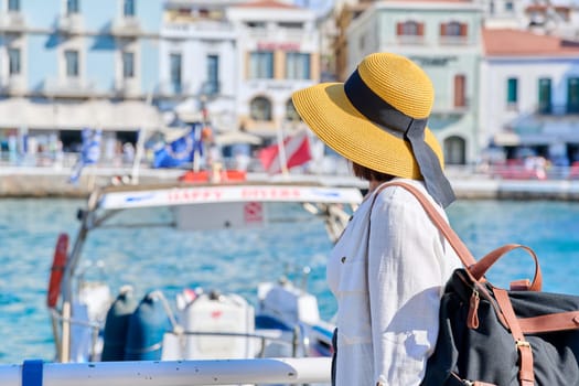 Beautiful mature woman in hat walking in seaside european city, copy space. Female tourist with backpack on the embankment on sunny summer day. Tourism, travel, vacation, people concept