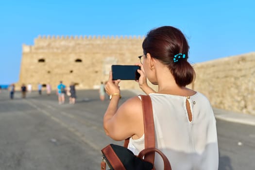 Woman tourist taking photo on smartphone, an ancient historical fortress on seashore. Tourism, travel, people, Greece Crete Heraklion