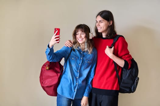 Couple of happy teenage students taking selfie using smartphone. Friends students with backpacks on the background of light wall. Youth, college, high school, education, young people concept