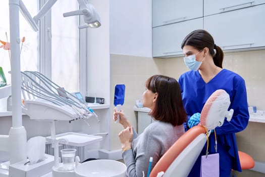 Dentist's office, woman patient looking at her teeth in the mirror. Doctor dentist near the dental chair with tools. Treatment, dental care, prosthetics, orthodontics, dentistry concept