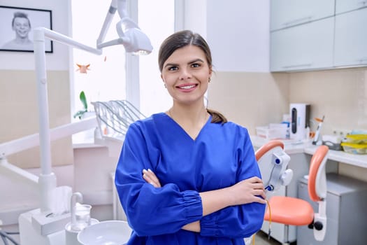 Portrait of confident female dentist in office looking at camera. Young female doctor in blue uniform with crossed arms. Dentistry, medicine, health care, profession, stomatology concept