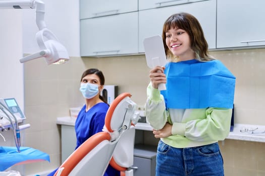 Teenage female looking at healthy teeth in mirror, in dental office, visit to dentist orthodontist. Treatment and care of teeth, teenagers, orthodontics, dentistry concept
