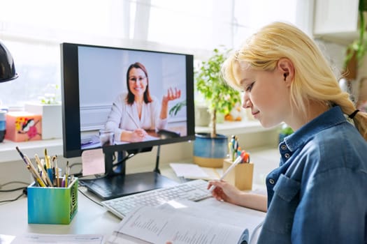 Teenage girl studying at home online using a computer. Virtual lesson, female teacher on the screen talking to a student, teaching remotely. E-learning, video conference, adolescence, high school concept