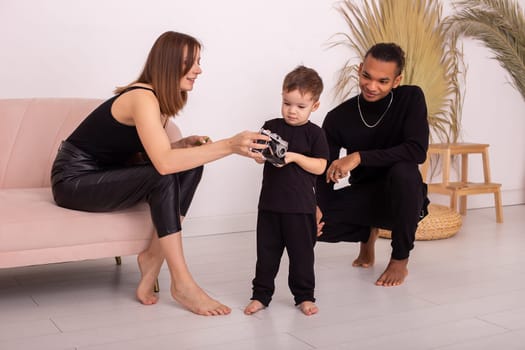 Happy multiracial family with little boy, in black clothes, in the light living room, playing , hold old camera