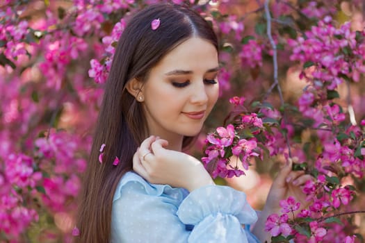 Portrait of beautiful brunette girl with pink petals in long hair stands near a pink blooming apple tree. Close up