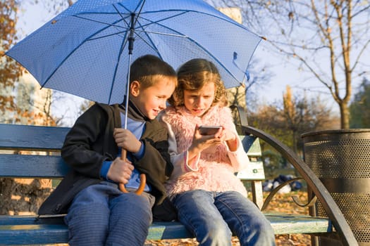 Outdoor portrait of two smiling children boy and girl, sitting under an umbrella on bench in autumn park, using smartphone, golden hour