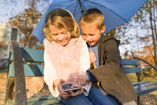 Outdoor portrait of two smiling children boy and girl, sitting under an umbrella on bench in autumn park, using smartphone, golden hour