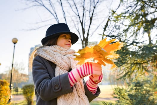 Autumn outdoor portrait of girl with yellow maple leaves, park background at golden hour, child in coat hat