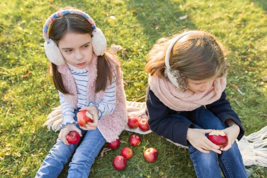 Children two girls eating red apples sitting in yellow autumn park, golden hour