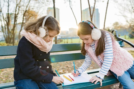 Two little pretty artists drawing with colored pencils, girls sitting on a bench in sunny autumn park, golden hour