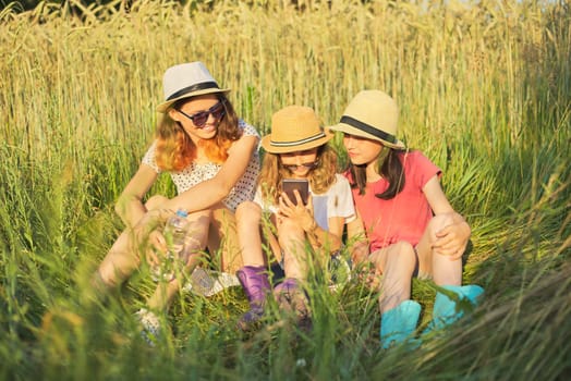 Portrait of three girls sitting and resting in grass, children talking, looking at smartphone laughing on sunny summer day, sunset in meadow