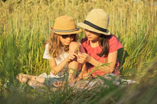 Two smiling girls sitting in grass, children talking and enjoying summer day at sunset