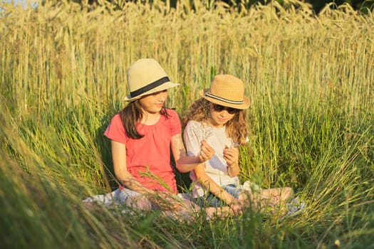 Two smiling girls sitting in grass, children talking and enjoying summer day, telling their secrets at sunset