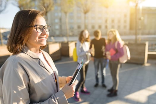 Portrait of mature smiling female teacher in glasses with clipboard, outdoor with a group of teenagers students, golden hour