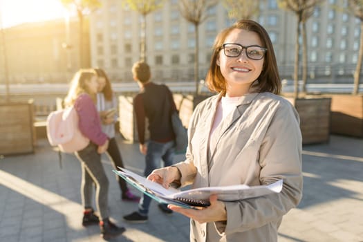 Portrait of mature smiling female teacher in glasses with clipboard, outdoor with a group of teenagers students, golden hour
