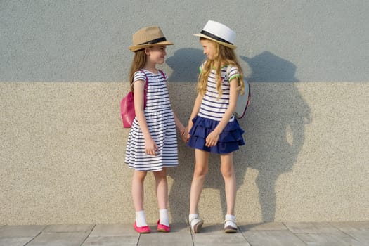 Outdoor summer portrait of two happy girl friends 7,8 years in profile talking and laughing. Girls in striped dresses, hats with backpack, background gray wall