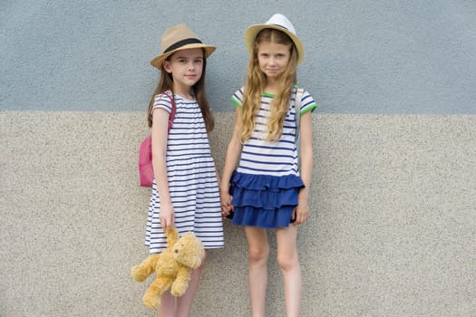Outdoor summer portrait of two happy girl friends 7,8 years holding hands. Girls in striped dresses, hats with backpack, background gray wall