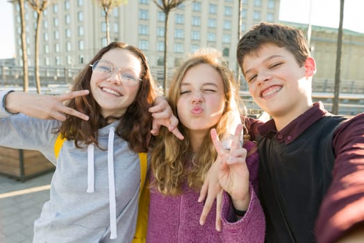 Portrait of friends teen boy and two girls smiling, making funny faces, showing victory sign in the street. City background, golden hour