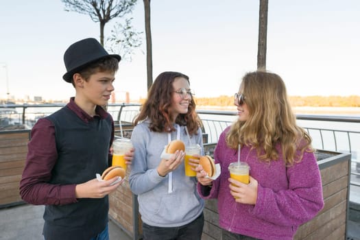 Teenagers eat street food, friends boy and two girls on city street with burgers and orange juice. City background, golden hour