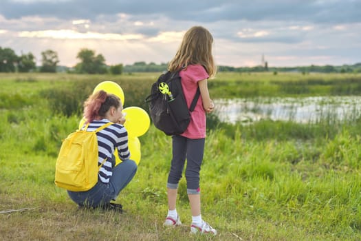 Two girls with balloons backpacks back in nature, children near the lake in the meadow, summer evening