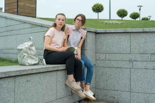 Outdoor portrait of two young beautiful girls students with backpacks, books. Girls talking, looking in book, urban background