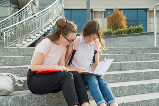Outdoor portrait of two young beautiful girls students with backpacks, books. Girls talking, looking in book, urban background