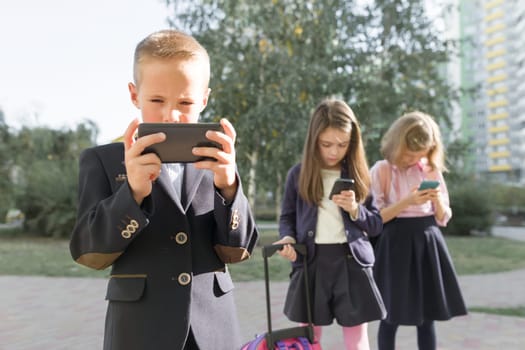 Group of children 7, 8 years old with mobile phones, schoolchildren with backpacks looking into smartphones, outdoor background. Education, friendship, technology and people concept