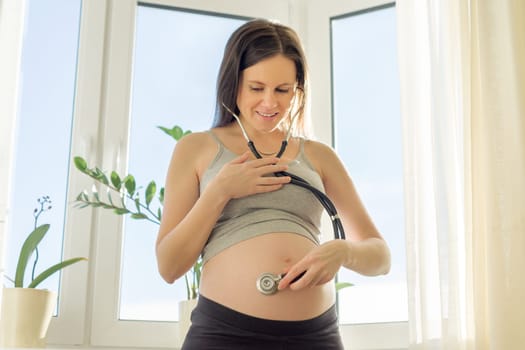 Beautiful young pregnant woman with stethoscope listening to her belly with baby at home near the window