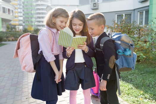 Outdoor portrait of smiling schoolchildren in elementary school. Group of kids with backpacks are having fun, talking, reading a book. Education, friendship, technology and people concept