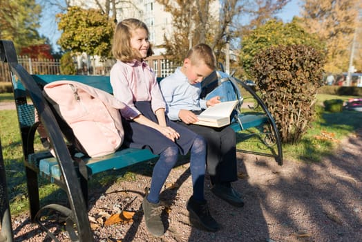 Little boy and girl schoolchildren reading book, sitting on bench, children with backpacks, bright sunny autumn day