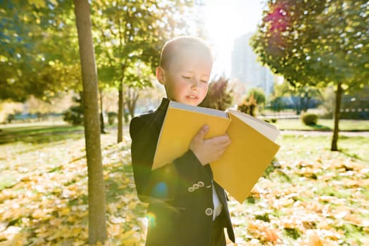 Outdoor autumn portrait of schoolboy reading book, background of yellow trees in the park, boy in jacket, golden hour, back to school