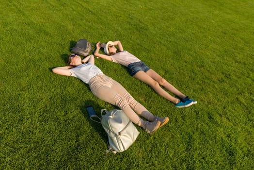 Two young female students with backpacks lying on green lawn grass. Top view, side view