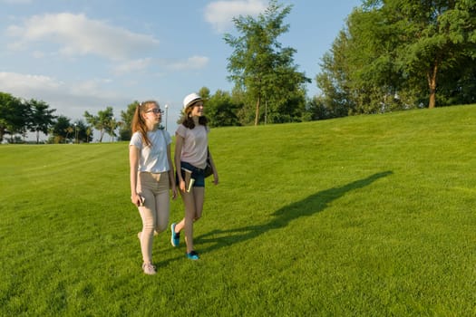 Two teen girls students with backpacks and books walking on green grass in the park, sunny day