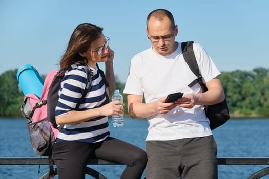 Outdoor mature couple using smartphone, man and woman talking walking in the park, people in sportswear with bottle of water, summer sunny evening near the river