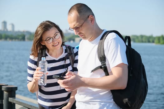 Outdoor mature couple using smartphone, man and woman talking walking in the park, people in sportswear with bottle of water, summer sunny evening near the river